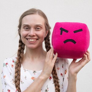 May include: A plush, magenta cube with a black, angry face. The cube is held by a person wearing a floral top. The background is a plain white wall. The cube is a fun, expressive item.