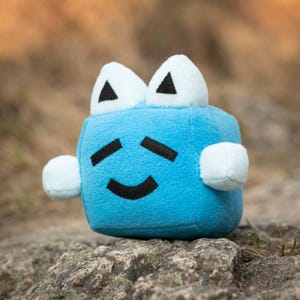 May include: A blue plush toy shaped like a cube with a smiling face and white ears. The toy is sitting on a rock.