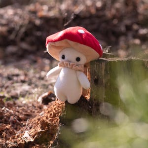 May include: A plush mushroom figure with a red cap dotted with white spots. The figure has a beige stem, a ruffled collar, and small arms. It is set against a blurred natural background, perched on a wooden stump.