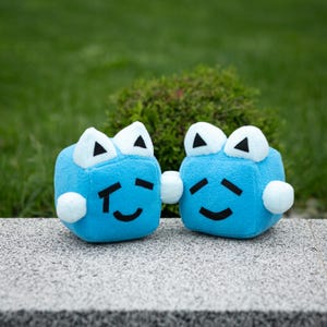 May include: Two plush blue cube toys with white triangle eyes and small white pom-pom arms. Each cube has a black curved mouth and a black line on each side of the mouth. The toys are sitting on a gray surface with a green background.
