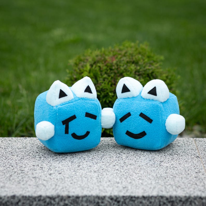 May include: Two plush blue cube toys with white triangle eyes and small white pom-pom arms. Each cube has a black curved mouth and a black line on each side of the mouth. The toys are sitting on a gray surface with a green background.