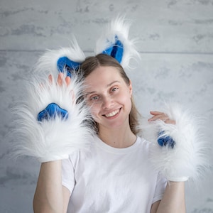 May include: White furry paw gloves and matching ear headband with blue accents. The gloves have blue paw pads. The ear headband has blue inner ears. The items are made of soft, fluffy material.