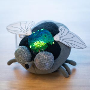 Könnte beinhalten: Ein Plüschspielzeug in Form einer Fliege mit großen, glitzernden Augen und einem Körper aus grauem, schwarzem und türkisfarbenem Stoff. Die Fliege hat weiße Papierflügel und einen Paillettenfleck auf dem Rücken. Das Spielzeug ist auf einer Holzoberfläche platziert.