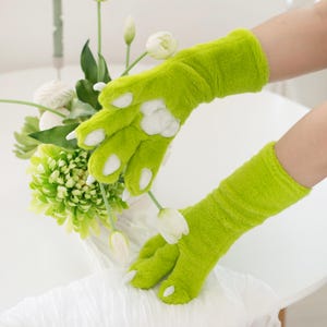 May include: Lime green plush gloves with white paw pads and claw-like tips. The gloves are designed to resemble a creature's paws. The gloves are shown near a white vase with green and white flowers.