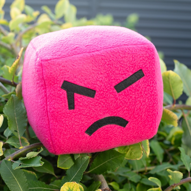 May include: A plush, cube-shaped toy in a vibrant pink color. The toy features a black, angry face design, with two angled lines for eyebrows and a curved line for the mouth. The toy is set against a backdrop of green leaves.