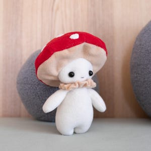 May include: A plush mushroom figure with a red cap dotted with white spots. The figure has a white body, small black eyes, and a ruffled beige collar. The mushroom is a whimsical, handmade item.