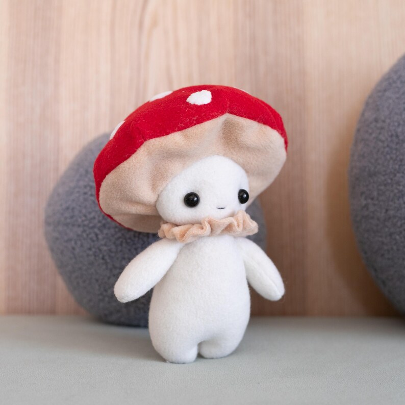 May include: A plush mushroom figure with a red cap dotted with white spots. The figure has a white body, small black eyes, and a ruffled beige collar. The mushroom is a whimsical, handmade item.