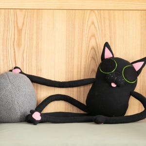 May include: A black cat plush toy with long arms and legs, pink accents on the ears and paws, and large green-outlined eyes. The cat is holding a gray, round plush object. The cat's tongue is sticking out.