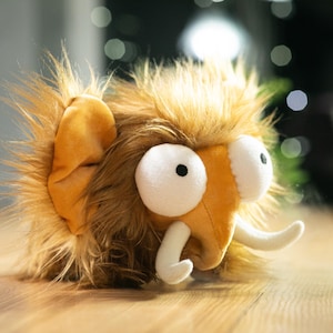 May include: A plush toy in the shape of a cartoon mammoth. The toy has large white eyes with black pupils, orange tusks, and a shaggy, brown and orange fur. The ears are orange.