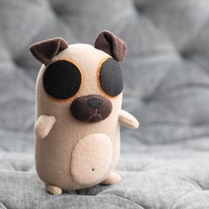 Może przedstawiać: Ręcznie robiona pluszowa zabawka w kształcie mopsa. Zabawka jest beżowa z dużymi czarnymi oczami obrysowanymi pomarańczem, brązowymi uszami i brązowym nosem. Mops ma małe ramiona i nogi i jest wykonany z miękkiej tkaniny.
