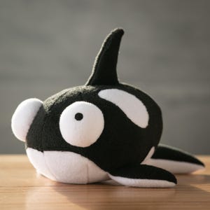 Puede incluir: Un peluche de orca en blanco y negro. El juguete tiene un cuerpo redondeado, una aleta dorsal y manchas blancas. La orca está sobre una superficie de madera. El juguete está diseñado para parecerse a una orca.