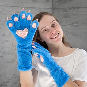 May include: Blue plush gloves with a paw design. The gloves have pink accents on the pads and claws, and a heart shape on the palm. The gloves are long-sleeved and made of a soft, furry material.