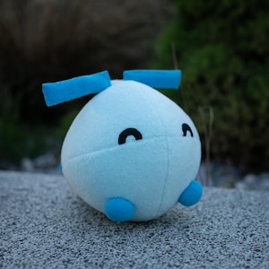 May include: A light blue plush toy with a rounded body, two blue antennae, and two small blue feet. The toy has black curved eyes and is made of soft fabric. The background is blurred, with green foliage and a gray surface.
