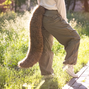 Può includere: Una lunga e folta coda in pelliccia sintetica color marrone caldo attaccata al retro di un pantalone cargo verde oliva. La coda presenta fibre dense e arruffate con varie tonalità di marrone che si estendono verso il basso dalla vita.