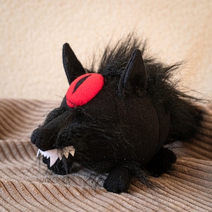 Puede incluir: Un peluche negro con forma de lobo, con un ojo rojo y colmillos blancos. El lobo tiene pelo negro alrededor de la cabeza y las orejas. El juguete está sobre una superficie marrón texturizada.