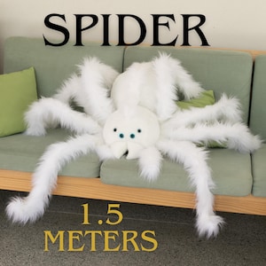 Pode incluir: Uma grande aranha de pelúcia branca com pernas longas e fofas e um corpo redondo. A aranha tem olhos azuis e mede aproximadamente 1,5 metros de comprimento. A palavra "SPIDER" está impressa em preto acima.