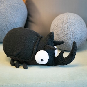 Puede incluir: Un juguete de peluche negro con forma de escarabajo, con ojos blancos grandes y un cuerno prominente. El juguete blando está sobre una superficie verde claro, con cojines esféricos grises en el fondo. El escarabajo tiene seis patas pequeñas.