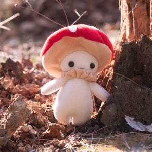 May include: A plush mushroom figure with a red cap and white polka dot. The mushroom has a beige stem and a small, smiling face with black eyes. It is standing in a natural outdoor setting.