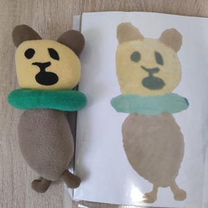 Pode incluir: Um urso de pel&uacute;cia marrom com rosto amarelo e olhos pretos usando um salva-vidas verde. Um desenho de um urso semelhante &eacute; mostrado em uma folha de papel branca.