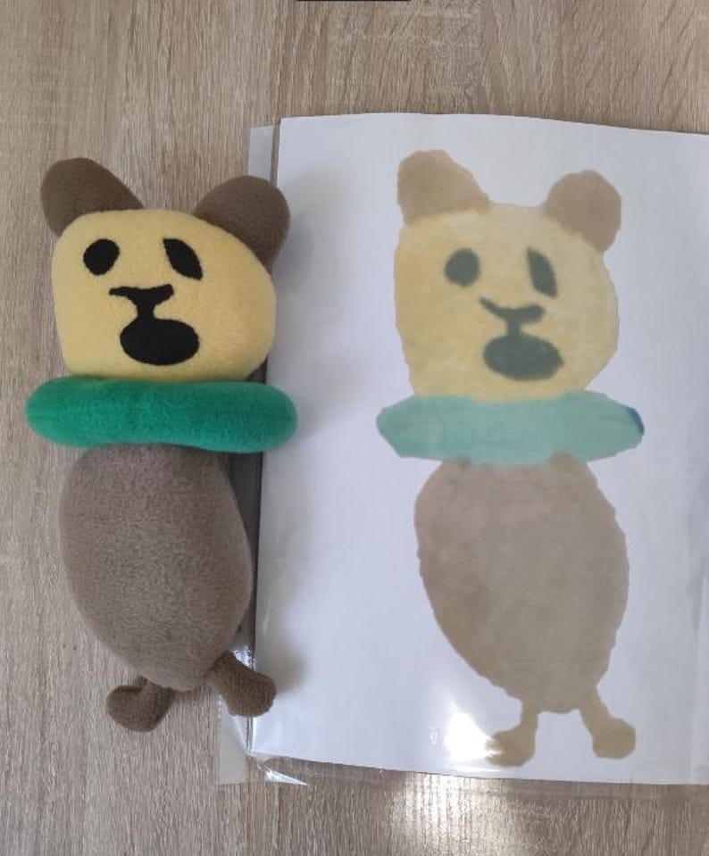 Pode incluir: Um urso de pel&uacute;cia marrom com rosto amarelo e olhos pretos usando um salva-vidas verde. Um desenho de um urso semelhante &eacute; mostrado em uma folha de papel branca.