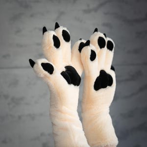 Puede incluir: Un par de guantes de felpa de color crema diseñados para parecerse a patas de animales. Cada guante presenta almohadillas negras y garras negras en las puntas de los dedos. Los guantes están hechos de un material suave y peludo.
