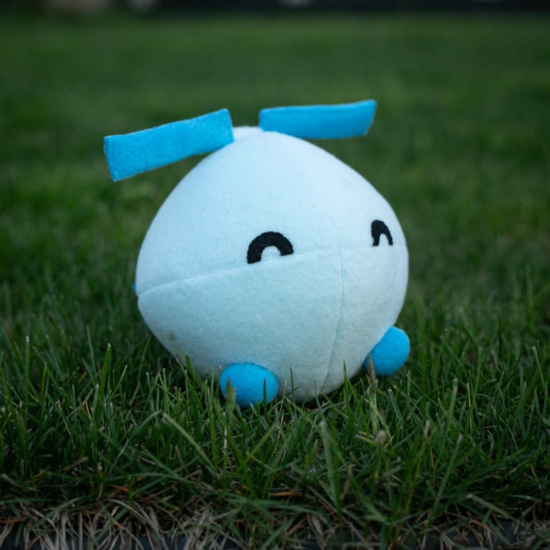 May include: A light blue plush toy with two blue rectangular appendages on top and two small blue feet. The toy has two black crescent-shaped eyes. The plush is sitting on a bed of green grass.