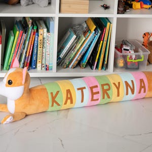 Puede incluir: Un cojín de peluche con forma de perro en tonos naranja, blanco y pastel. El cojín cilíndrico lleva el nombre "KATERYNA" cosido con letras marrones. El perro tiene orejas caídas y patas extendidas.
