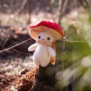 May include: A plush mushroom figure with a red cap dotted with white spots and a beige underside. The figure has a white body, small arms, and a ruffled beige collar. It is set against a blurred natural background.