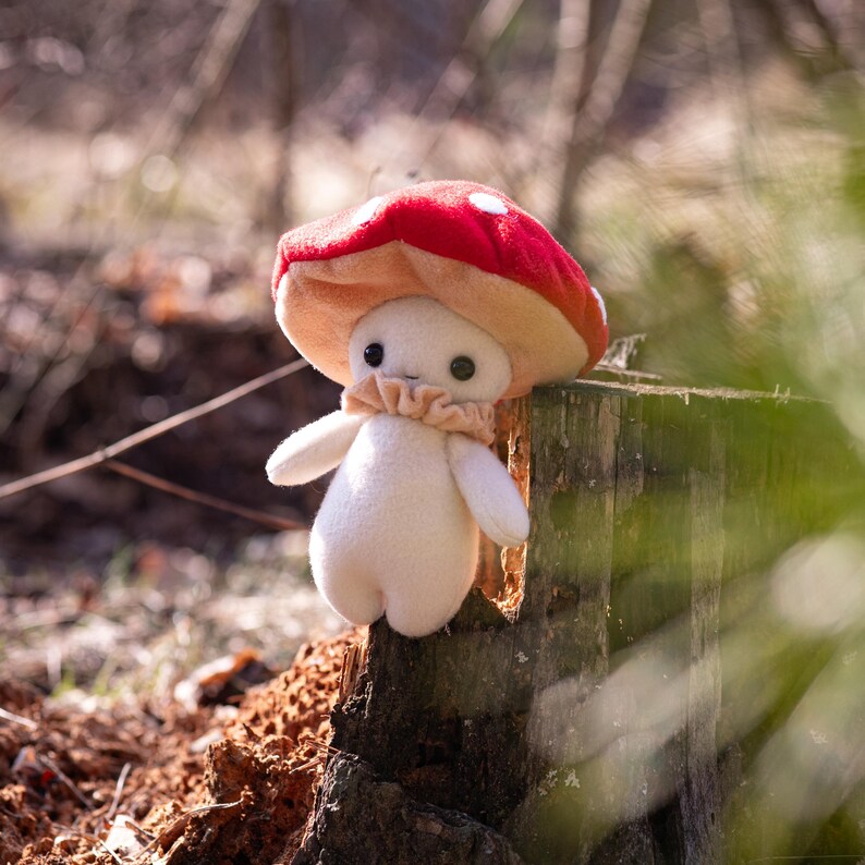 May include: A plush mushroom figure with a red cap dotted with white spots and a beige underside. The figure has a white body, small arms, and a ruffled beige collar. It is set against a blurred natural background.