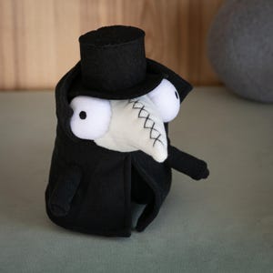 Peut inclure: Poupée artisanale en peluche représentant un médecin de la peste. La poupée est principalement noire, avec un masque blanc en forme de bec et de grands yeux blancs. Elle porte un haut-de-forme et un long manteau, pour une apparence gothique fantaisiste.