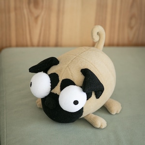 May include: A plush pug toy featuring a tan rounded body, a black face mask, and two oversized white circular eyes with small black pupils. The stuffed animal has small black ears, four short legs, and a curled tail extending upward from the back.