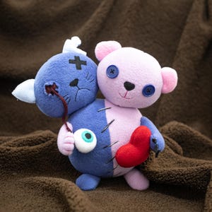 May include: A two-headed plush toy, one side a blue cat with a cross and exposed eye, the other a pink bear with button eyes. A red heart is sewn on the front. The toy is made of soft fabric. The toy is designed with a quirky, handmade aesthetic.