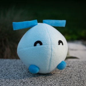 May include: A light blue plush toy with a rounded body, two blue rectangular appendages, and two small blue feet. The toy has two black curved eyes. The plush is made of soft material and is sitting on a gray surface.