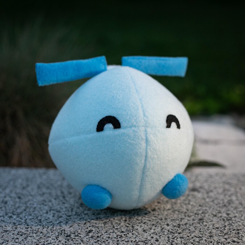May include: A light blue plush toy with a rounded body, two blue rectangular appendages, and two small blue feet. The toy has two black curved eyes. The plush is made of soft material and is sitting on a gray surface.