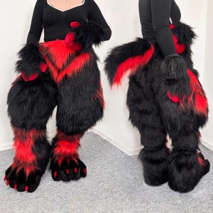 Op de afbeelding: Een volledig fursuit-kostuum van lang zwart en rood imitatiebont. De set bevat digitigrade beenvulling, grote pootvormige voeten met zwarte klauwen en rode accenten, bijpassende handpoten en een lange, dikke, borstelige staart met rood bont.