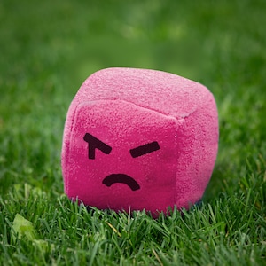 May include: A pink, cube-shaped plush toy with an angry face drawn on it. The toy is sitting on a bed of green grass. The face has black eyes and a curved mouth.