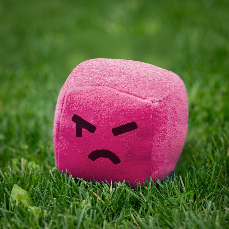 May include: A pink, cube-shaped plush toy with an angry face drawn on it. The toy is sitting on a bed of green grass. The face has black eyes and a curved mouth.