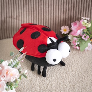 Peut inclure: Une peluche coccinelle avec un dos rouge vif orné de points noirs, un corps noir et deux grands yeux blancs ronds avec de petites pupilles noires. L'insecte en peluche possède de fines pattes noires et des antennes sur la tête.