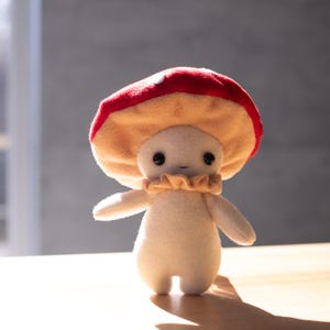 May include: A plush mushroom figure with a red and tan cap, a cream-colored body, and small black eyes. The figure has a ruffled collar and outstretched arms, standing on a wooden surface. The background is a blurred gray.