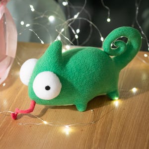 May include: A plush green chameleon toy with large white eyes and a pink tongue. The stuffed animal has a curled tail and is made of a soft, textured fabric. The toy is designed to resemble a cartoon chameleon.