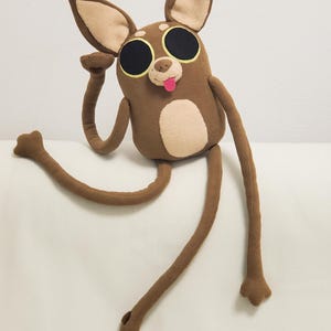 Puede incluir: Un peluche marrón con ojos negros grandes, una lengua rosa y extremidades largas y delgadas. El juguete tiene orejas internas de color canela y una mancha en el vientre de color canela. Un brazo está levantado y el otro extendido.