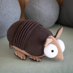 May include: A plush armadillo toy in shades of brown and tan, with large white eyes and small ears. The toy has a ridged back and is positioned on a green surface, with gray pillows in the background.