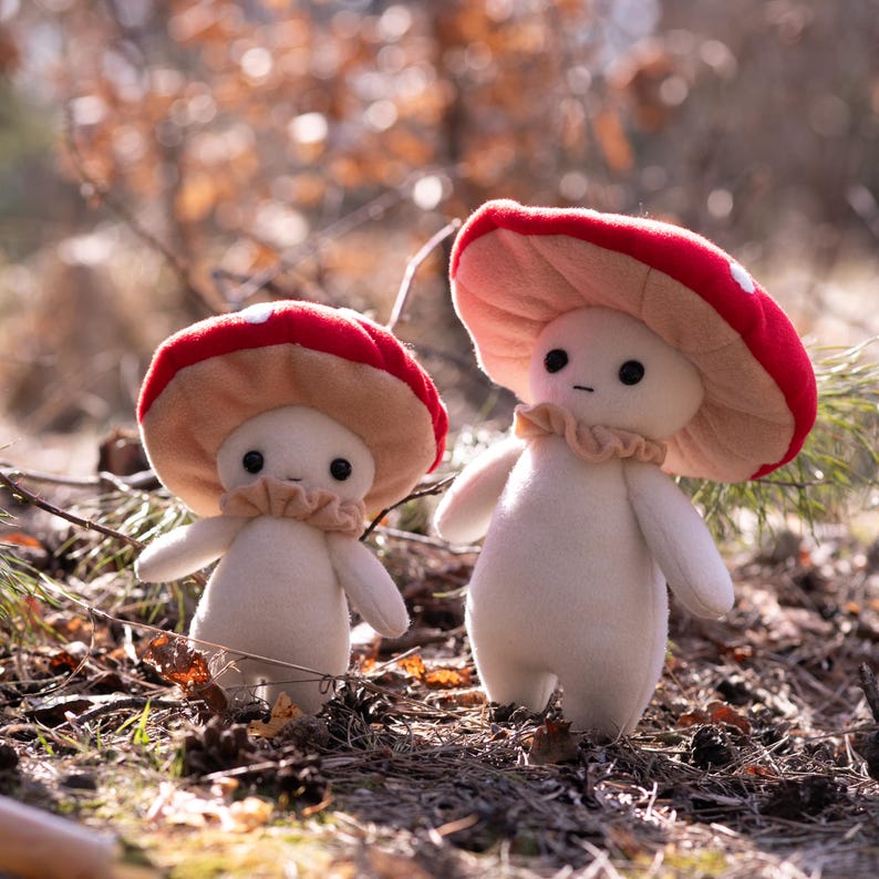 May include: Two plush mushroom figures with red and white spotted caps and beige frilly collars. The figures have white bodies and small black eyes. They are standing in a natural outdoor setting with leaves and pine needles.