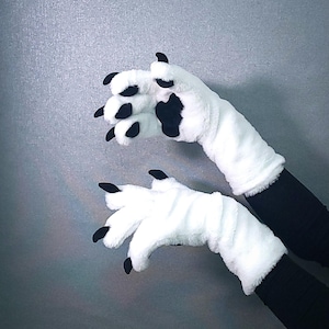 Puede incluir: Un par de guantes de felpa blancos con garras y almohadillas negras. Los guantes están diseñados para parecerse a las patas de animales, con largas garras negras curvadas que se extienden desde las puntas de los dedos. Los guantes se usan sobre mangas negras.