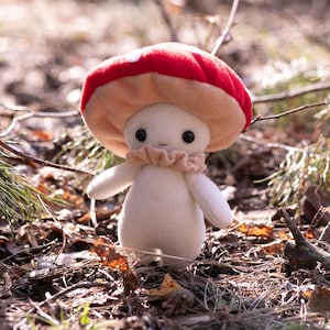 May include: A plush mushroom figure with a red and white spotted cap, a beige frilly collar, and a cream-colored body. The mushroom has black button eyes and small arms. The figure is standing in a natural outdoor setting.