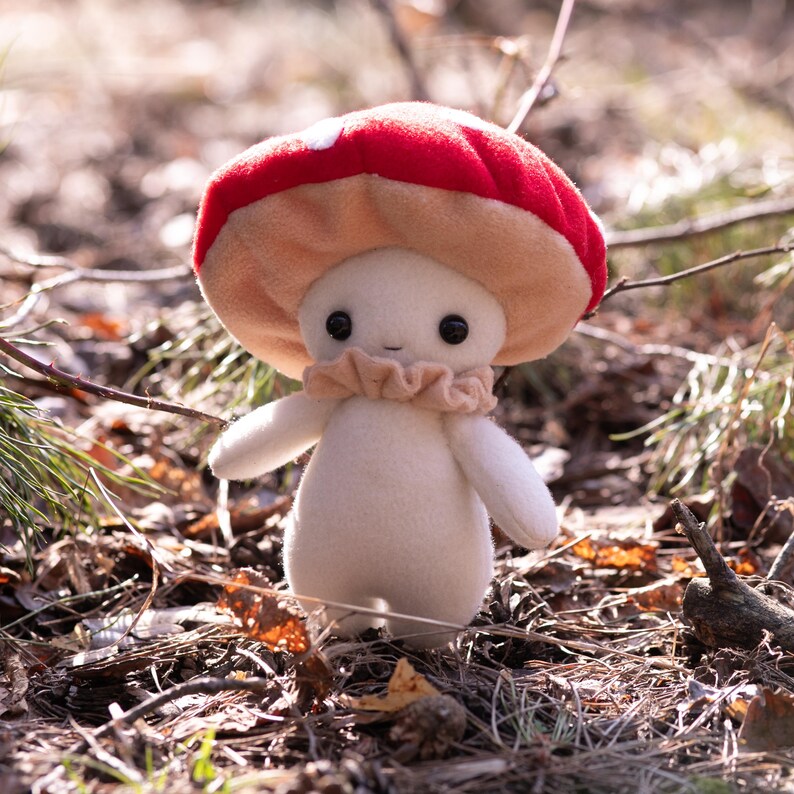 May include: A plush mushroom figure with a red and white spotted cap, a beige frilly collar, and a cream-colored body. The mushroom has black button eyes and small arms. The figure is standing in a natural outdoor setting.