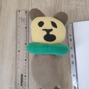 Pode incluir: Um brinquedo de pel&uacute;cia marrom e amarelo em forma de urso usando um salva-vidas verde. O urso tem um nariz e olhos pretos.