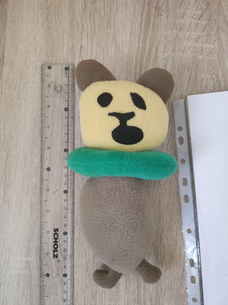 Pode incluir: Um brinquedo de pel&uacute;cia marrom e amarelo em forma de urso usando um salva-vidas verde. O urso tem um nariz e olhos pretos.