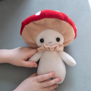 May include: A plush mushroom doll with a red cap featuring white spots, a tan underside, and a beige body. The doll has black button eyes and a ruffled collar. The doll is being held by a person.