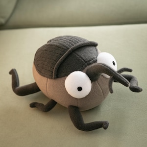 May include: A plush toy beetle with a gray segmented shell, large white eyes with black pupils, and gray appendages. The body is a light brown color, and the toy is resting on a green surface.